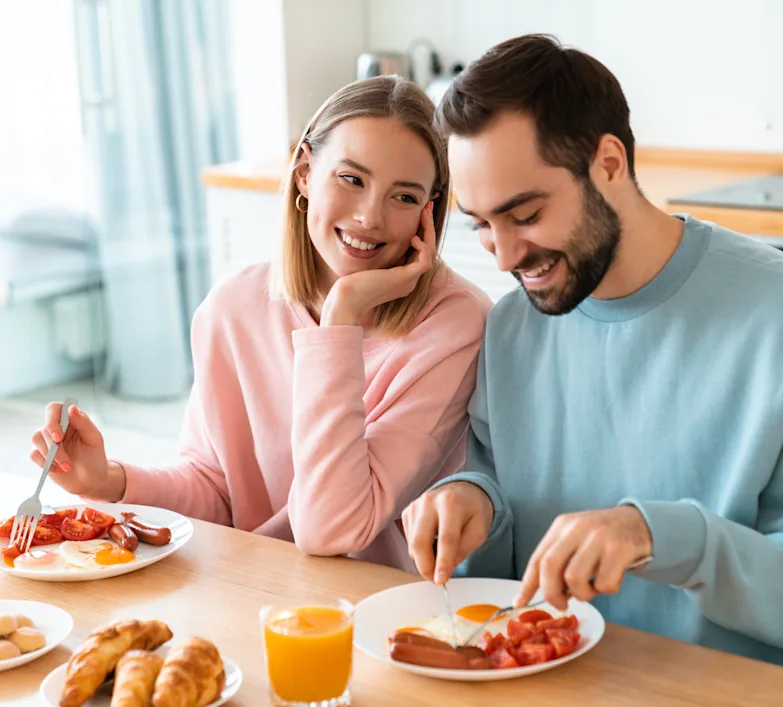Couple enjoying some breakfast together. The woman is looking lovingly at the man while he cuts the sausage on his plate.