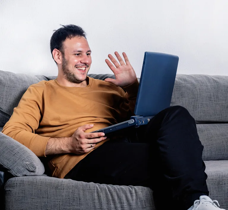 Man in a yellow, long-sleeved shirt waving at his computer while sitting on his gray couch