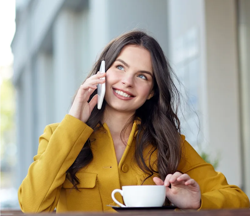 Woman in a yellow jacket calling on the phone. She has a white coffee cup in front of her.